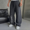 A person wearing loose, wide-leg gray sweatpants with a white script logo on the lower left leg, paired with a white t-shirt and white sneakers. The photo is taken in an indoor setting with a gray textured wall, a black leather sofa, and a gray floor.