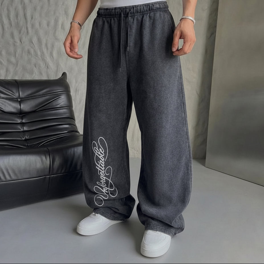 A person wearing loose, wide-leg gray sweatpants with a white script logo on the lower left leg, paired with a white t-shirt and white sneakers. The photo is taken in an indoor setting with a gray textured wall, a black leather sofa, and a gray floor.