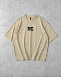 A beige short-sleeve t-shirt with a black 'DC' logo printed on the chest, laid flat on a gray tiled background.