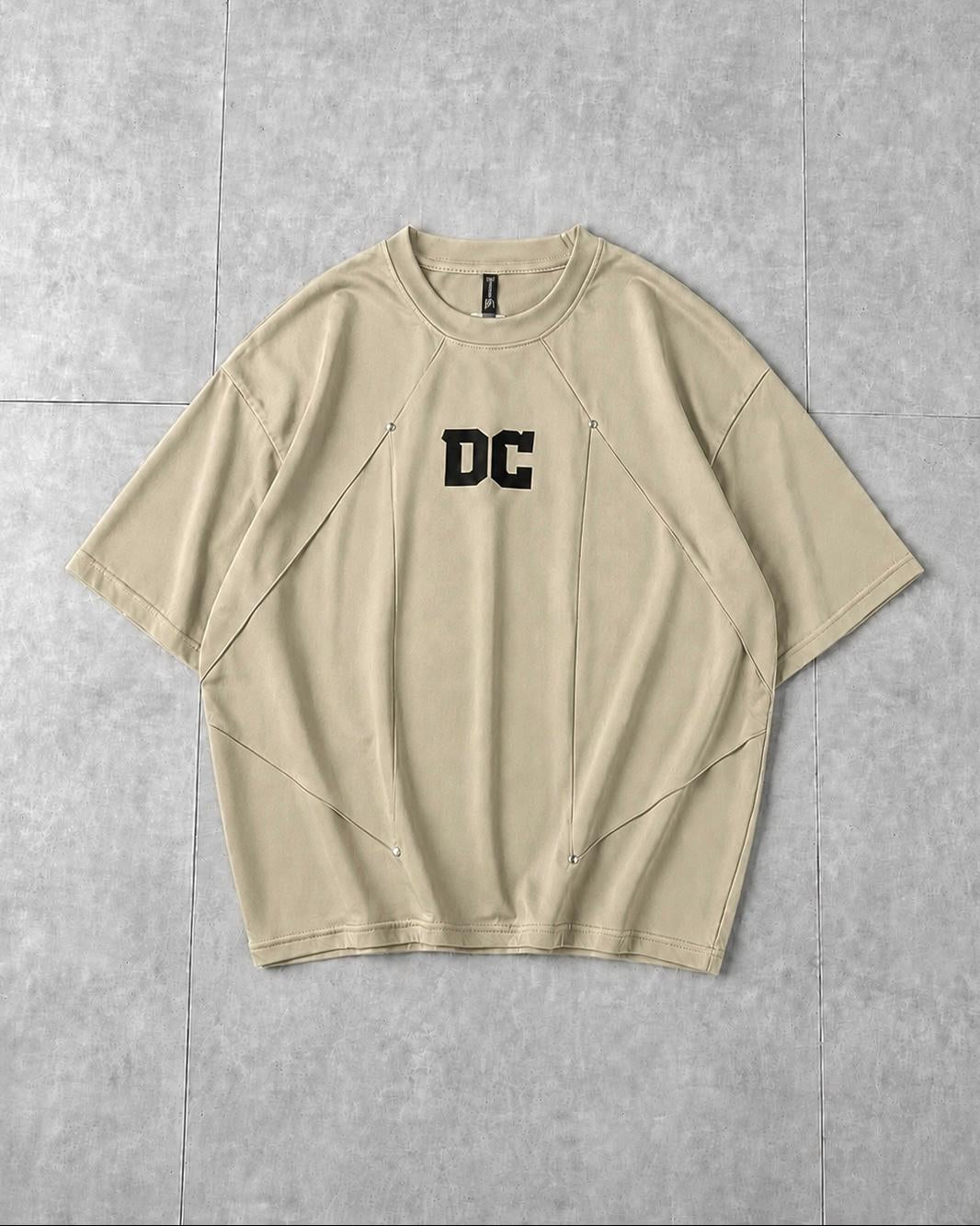 A beige short-sleeve t-shirt with a black 'DC' logo printed on the chest, laid flat on a gray tiled background.