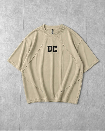 A beige short-sleeve t-shirt with a black 'DC' logo printed on the chest, laid flat on a gray tiled background.