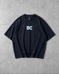 A black short-sleeve crewneck t-shirt with a white 'DC' logo printed on the chest, laid flat on a gray tiled background.