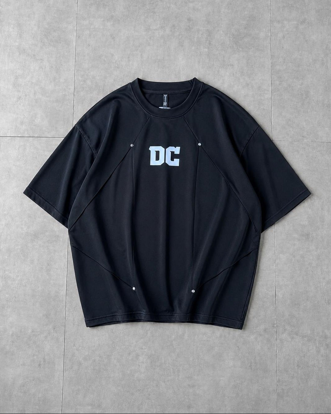 A black short-sleeve crewneck t-shirt with a white 'DC' logo printed on the chest, laid flat on a gray tiled background.