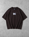 A dark brown short-sleeve t-shirt with a white 'DC' graphic on the chest, laid flat on a gray tiled surface.