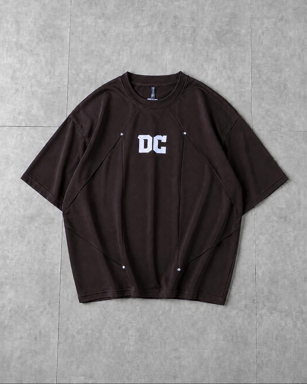 A dark brown short-sleeve t-shirt with a white 'DC' graphic on the chest, laid flat on a gray tiled surface.