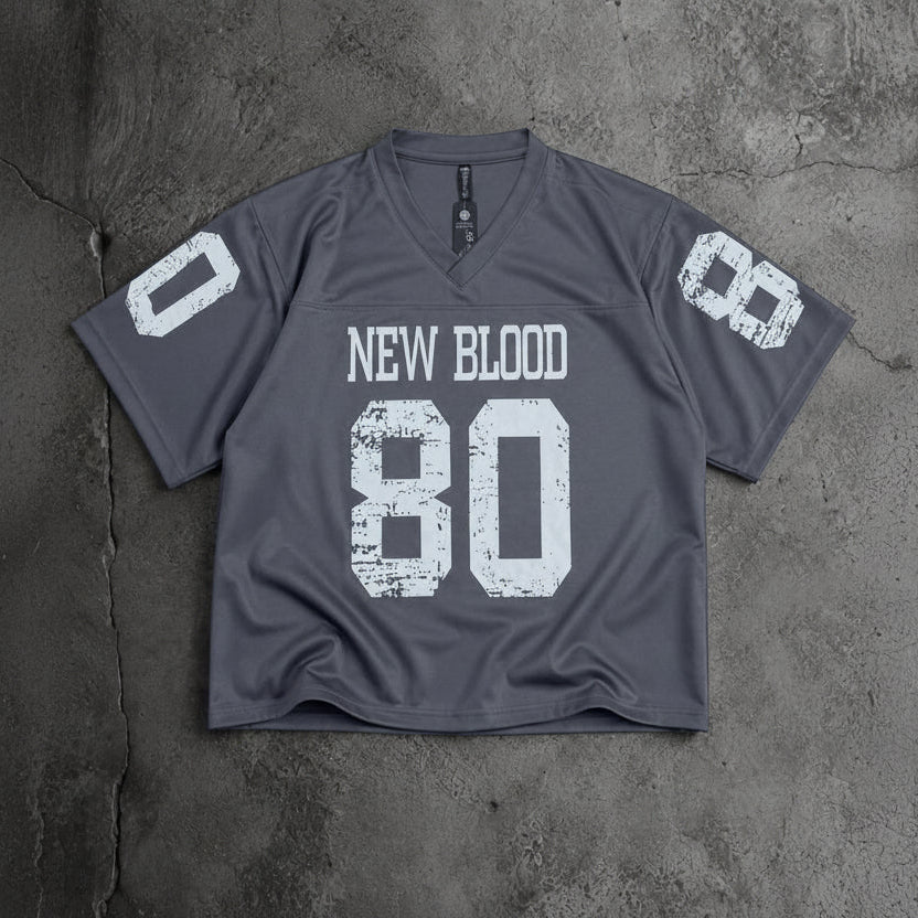 A dark gray v-neck athletic jersey with distressed white text reading 'NEW BLOOD' and the number '80' on the front, plus matching white numbers on the sleeves, laid flat on a textured gray concrete surface.