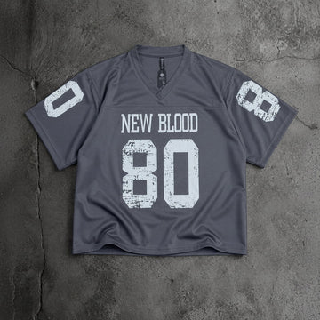 A dark gray v-neck athletic jersey with distressed white text reading 'NEW BLOOD' and the number '80' on the front, plus matching white numbers on the sleeves, laid flat on a textured gray concrete surface.